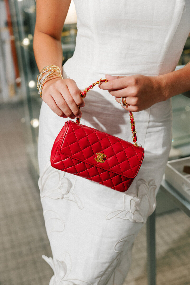 5 Formalwear Trends You'll See Everywhere This Summer - 11 Woman in a white dress holding a small red quilted handbag with a gold clasp and chain handle, showcasing Birmingham formalwear trends.