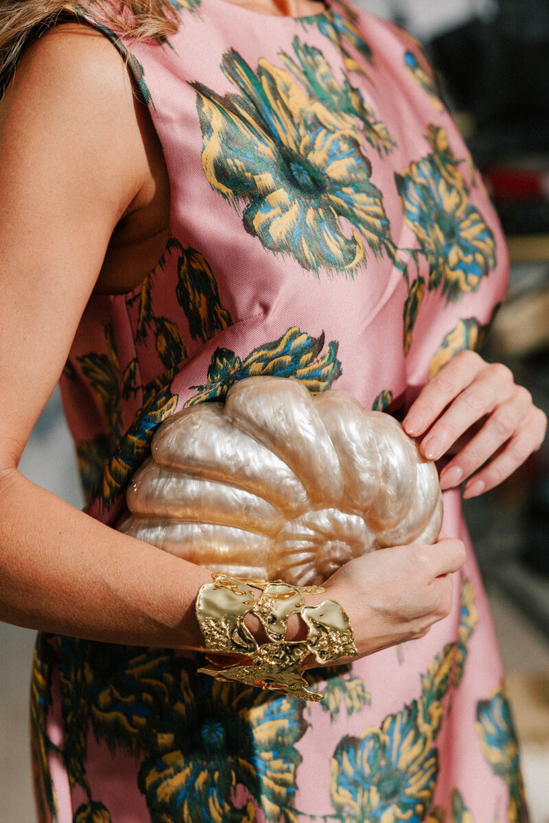 5 Formalwear Trends You'll See Everywhere This Summer - 13 A person in a pink and green floral dress holds a shell-shaped pearl clutch and wears a large gold bracelet β an elegant nod to Gus Mayer and the latest Birmingham formalwear trends.