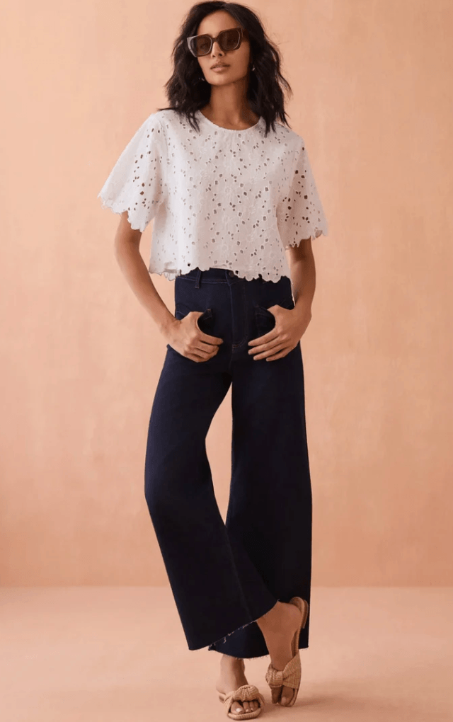 22 Birmingham FINDS for June: Breezy Dresses, Entertaining & Gifts for Dad - 6 A woman wearing sunglasses, a white eyelet short-sleeve crop top, high-waisted wide-leg dark jeans, and beige heeled sandals stands against a peach background.