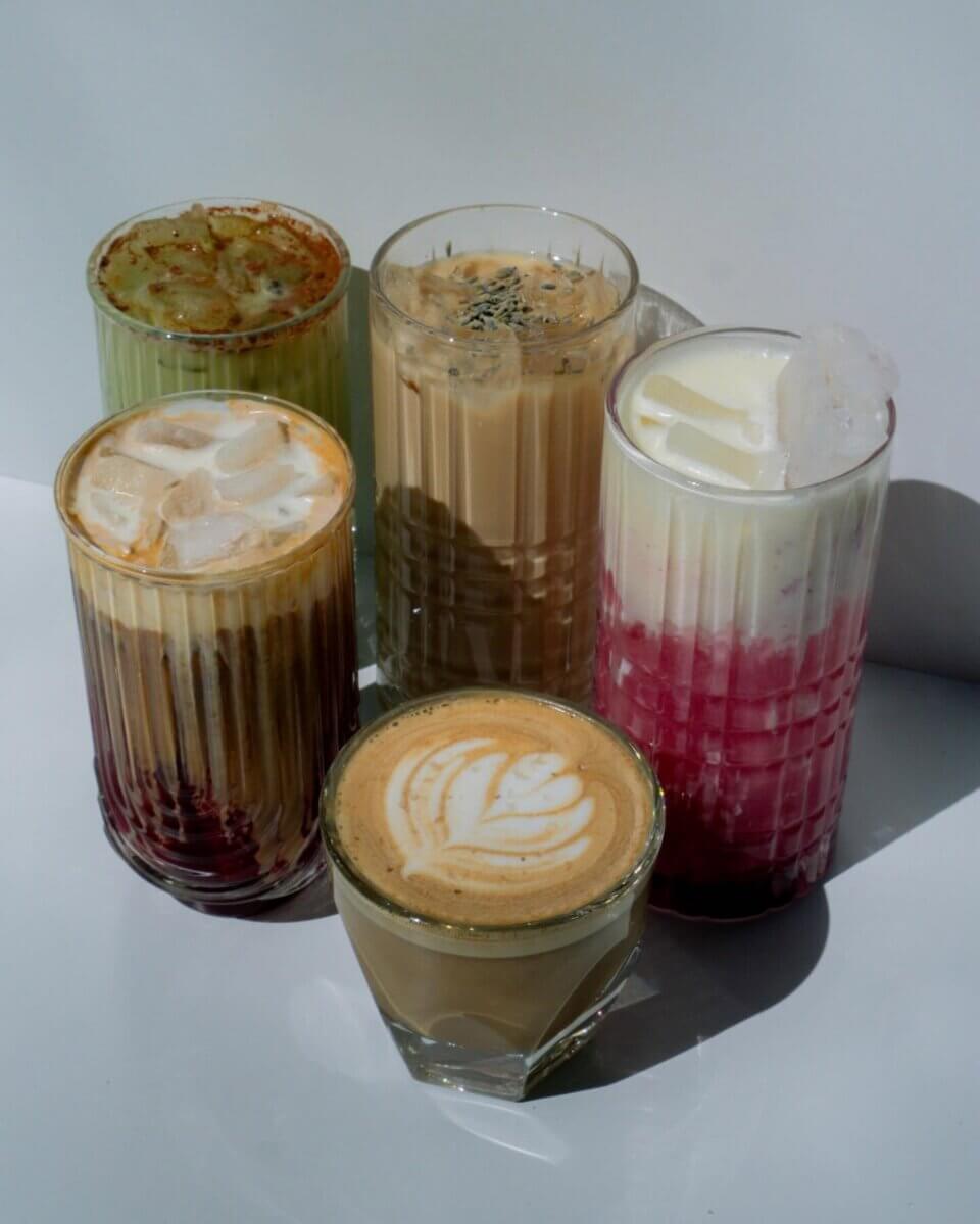 13 New Birmingham Restaurants to Try This Month - 3 Five glasses of colorful iced and hot drinks, including one with latte art, arranged on a white surface in bright lighting—a perfect snapshot of the creative offerings at new restaurants Birmingham has to offer.