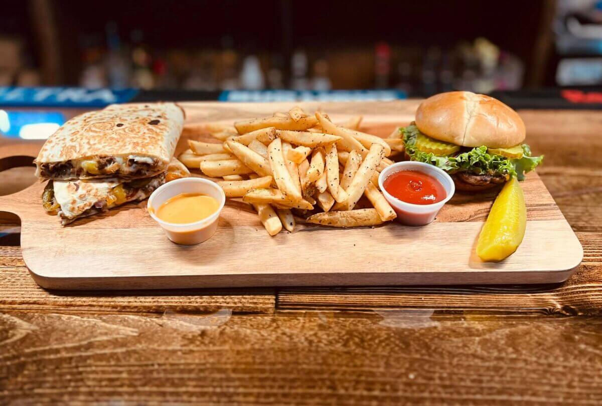 13 New Birmingham Restaurants to Try This Month - 5 A wooden board with a quesadilla, French fries, a hamburger with lettuce and tomato, two dipping sauces, and a pickle spear—perfect for sampling flavors at new restaurants Birmingham has to offer.