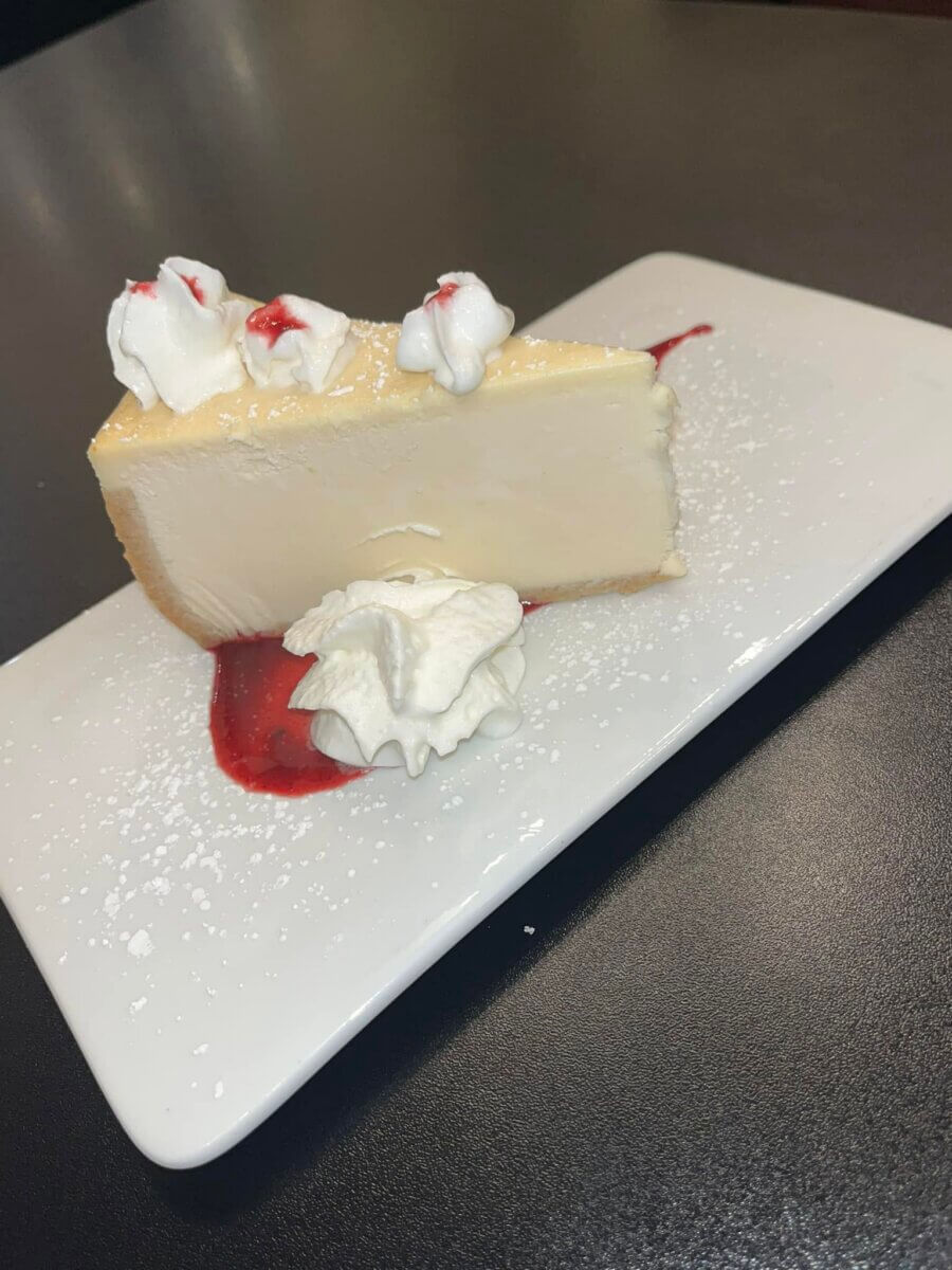 13 New Birmingham Restaurants to Try This Month - 6 A slice of cheesecake with whipped cream and red sauce on a white rectangular plate, dusted with powdered sugar.