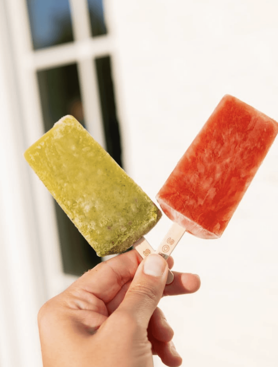 13 New Birmingham Restaurants to Try This Month - 12 A hand holding two popsicles, one green and one red, with a blurred white wall and window in the background.