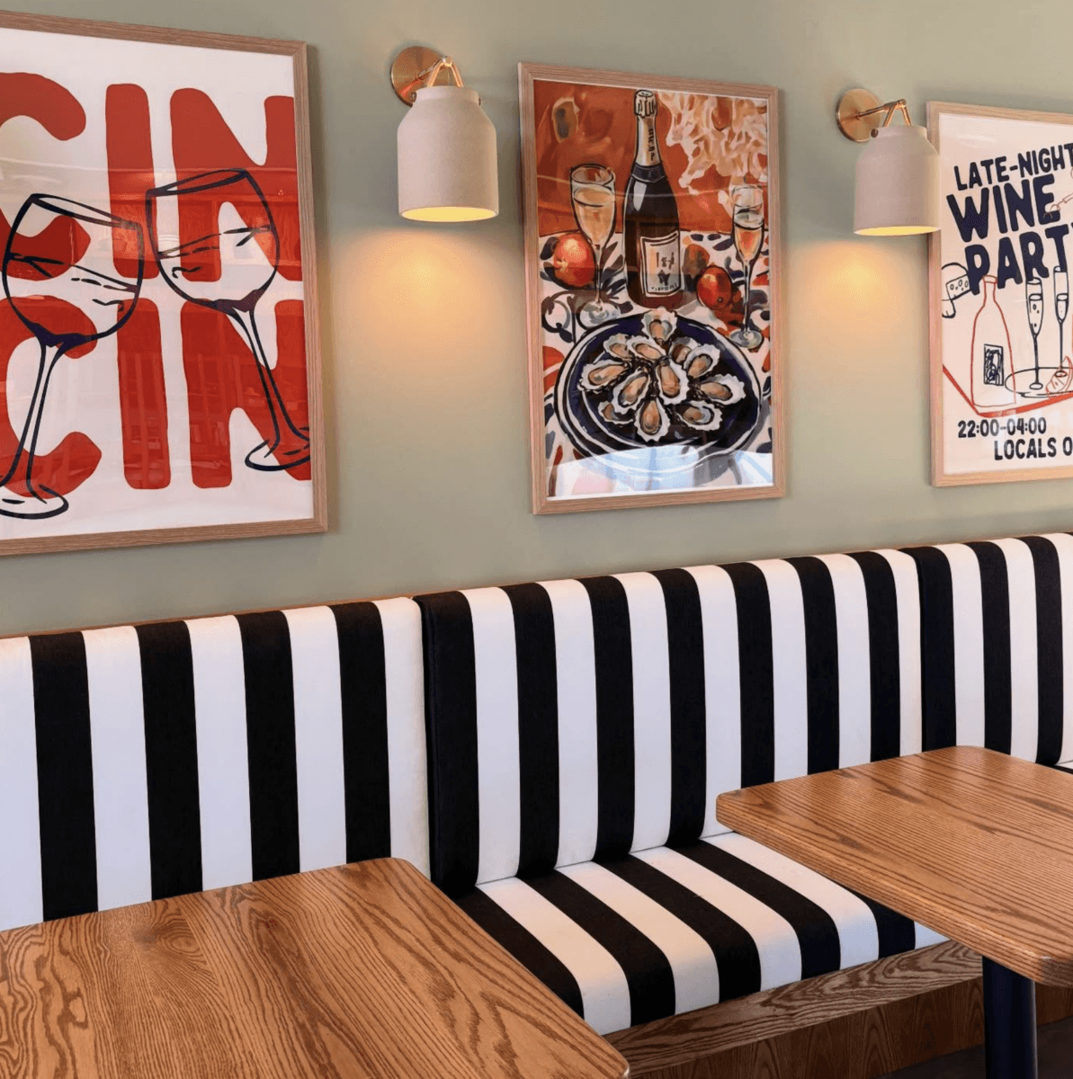13 New Birmingham Restaurants to Try This Month - 14 A restaurant booth in Birmingham with black and white striped seating, wooden tables, wall lamps, and colorful framed art posters on a light green wall—a stylish spot among the city’s new restaurants.