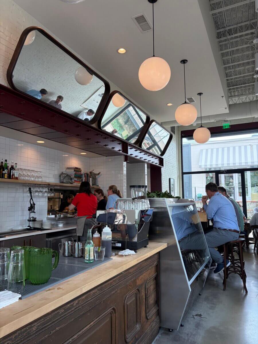 Modern café interior at Pepper Place, with customers seated at the counter and tables, overhead globe lights, and large mirrors on the wall reflecting the space—a perfect lunch spot.