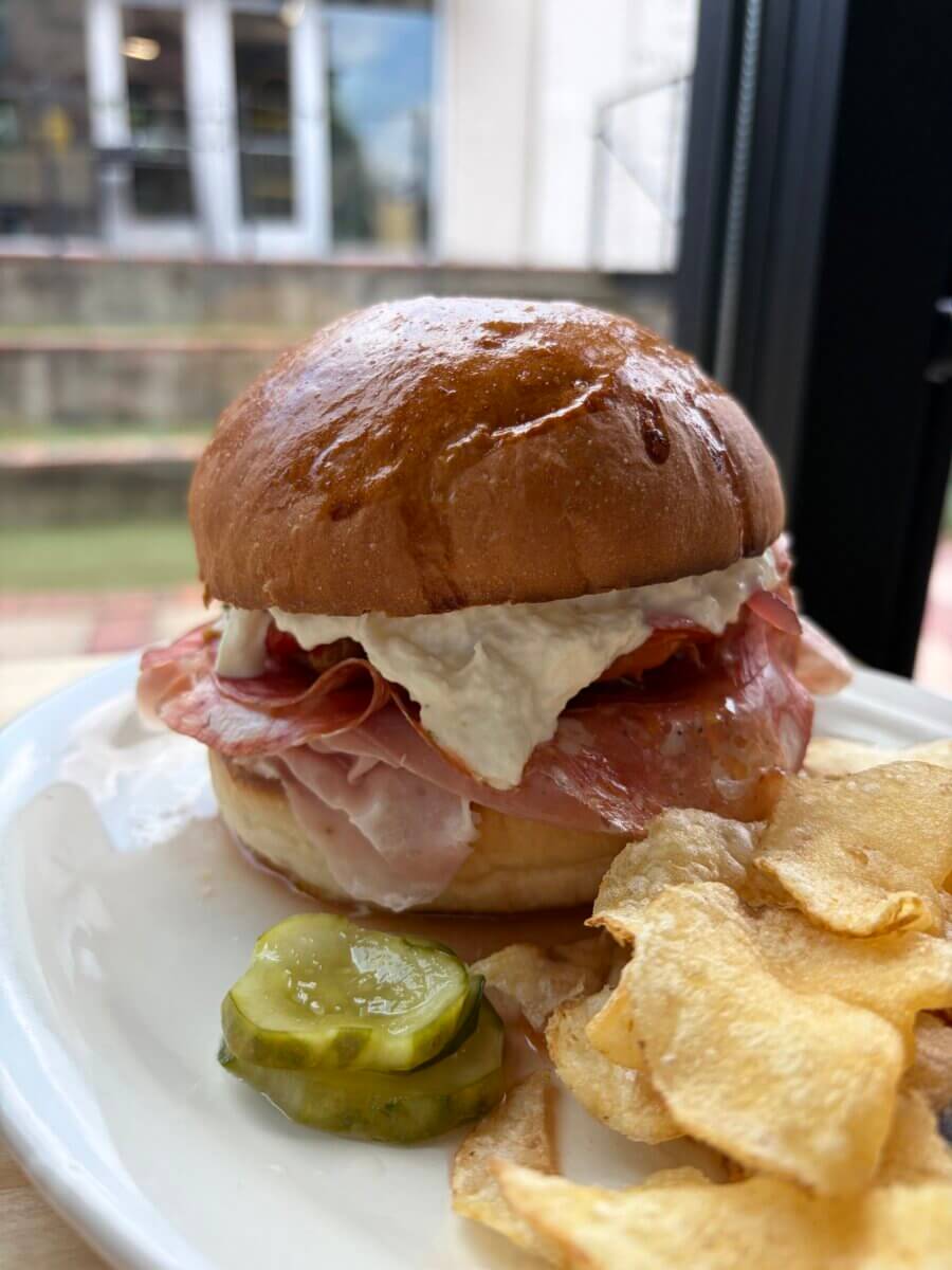A sandwich from The Mustard Club with cured meats and creamy spread on a brioche bun, served with pickles and potato chips.