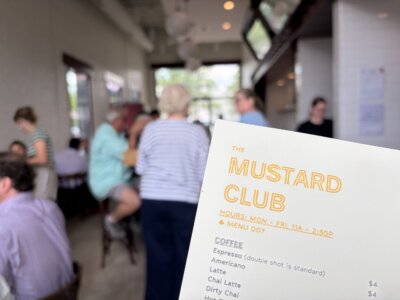 A close-up of The Mustard Club menu with prices, held in a busy, casual café at Pepper Place, filled with seated and standing patrons—an inviting lunch spot bustling with energy.