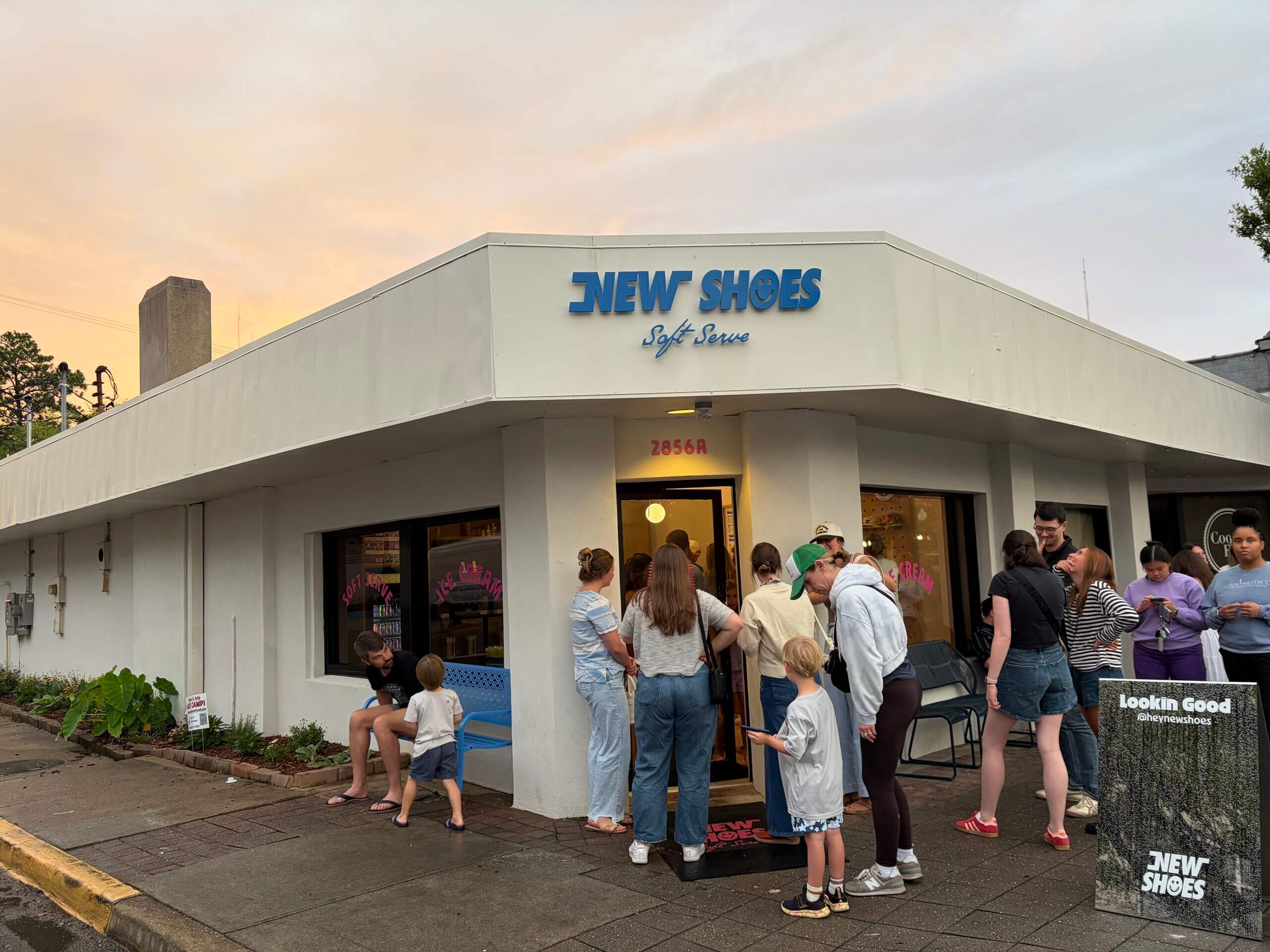A group of people, including children and adults, gather outside New Shoes Soft Serve in Homewood at dusk.