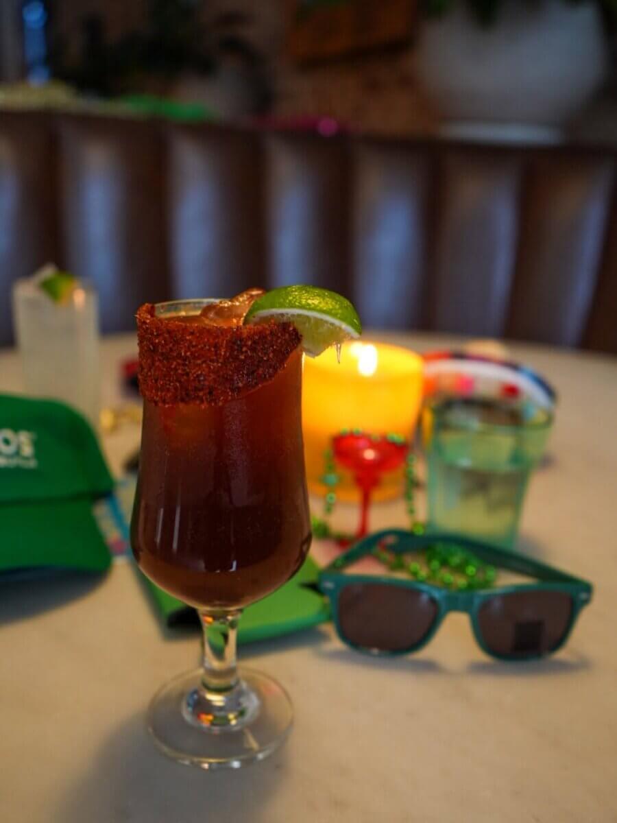 15+ Birmingham Restaurants Open on July 4th - 20 A cocktail with a chili-rimmed glass and lime wedge sits on a table at one of Birmingham restaurants, with green sunglasses, a candle, and drinks in the background—perfect for celebrating July 4th.
