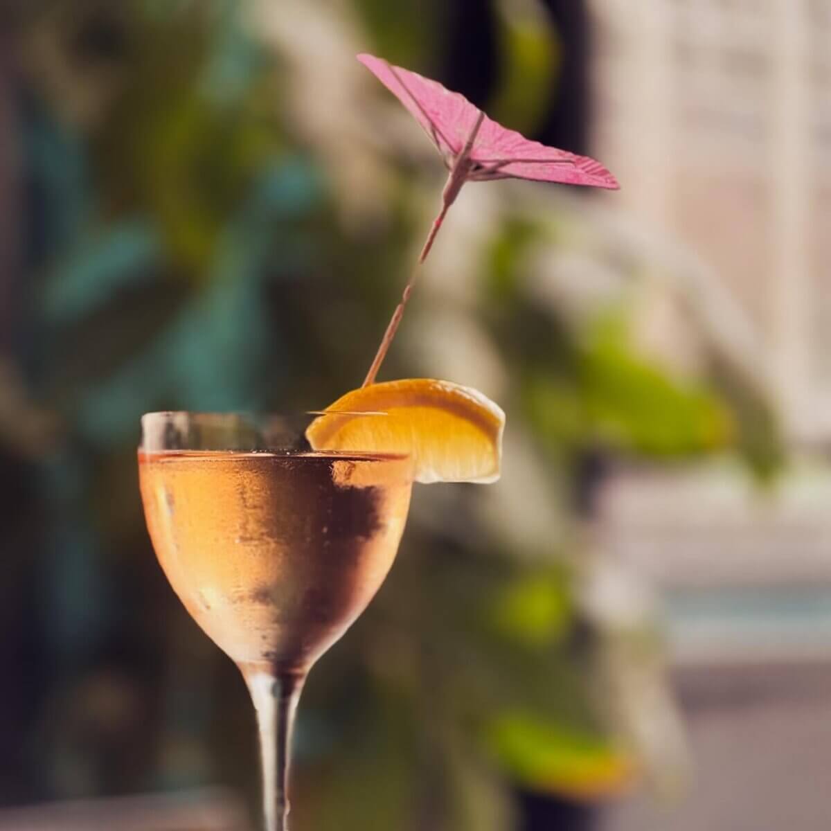 15+ Birmingham Restaurants Open on July 4th - 13 A stemmed glass of a golden drink with condensation, garnished with an orange slice and a pink paper umbrella, set against a blurred outdoor background—perfect for July 4th celebrations at Birmingham restaurants open this summer.