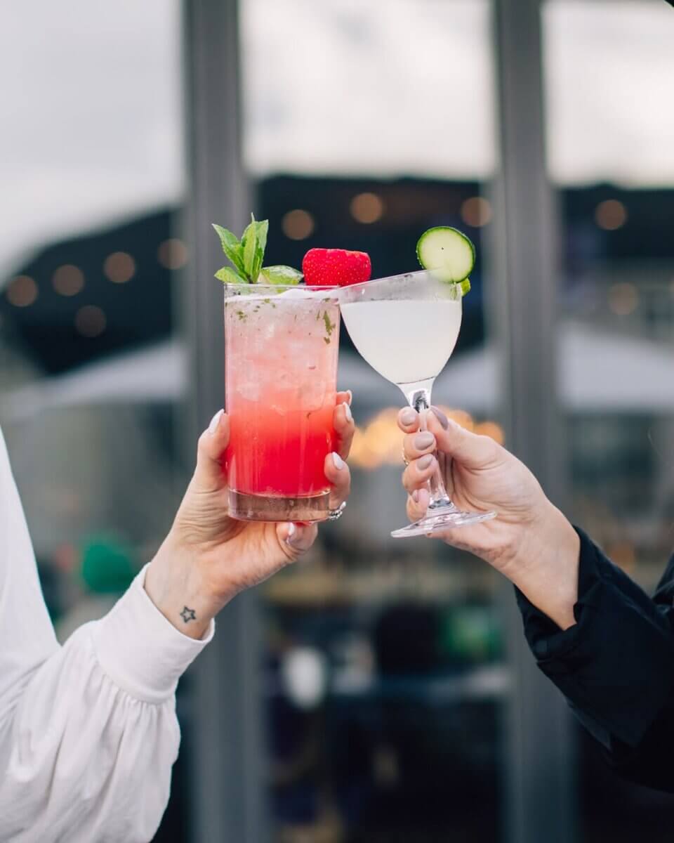 15+ Birmingham Restaurants Open on July 4th - 11 Two people clink glasses at one of the newest Birmingham restaurants—one with a pink cocktail garnished with mint and raspberries, the other with a clear drink and cucumber slice—celebrating an open, lively July 4th evening.