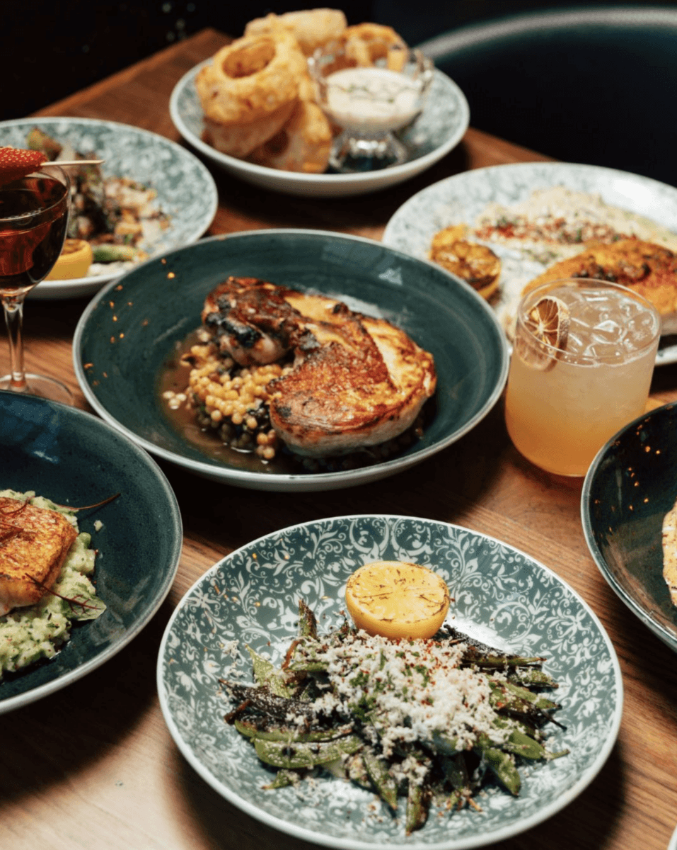 15+ Birmingham Restaurants Open on July 4th - 10 A festive table set with plates of cooked vegetables, seared chicken, grilled fish, onion rings, and drinks garnished with fruit and citrus on patterned green dinnerware—perfect for a July 4th celebration at Birmingham restaurants open for the holiday.