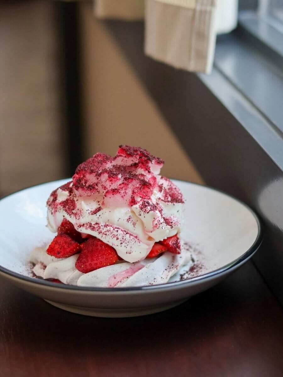 15+ Birmingham Restaurants Open on July 4th - 9 A dessert with whipped cream and sliced strawberries on a white plate, topped with red powder, placed near a window—perfect for enjoying at Birmingham restaurants open July 4th.