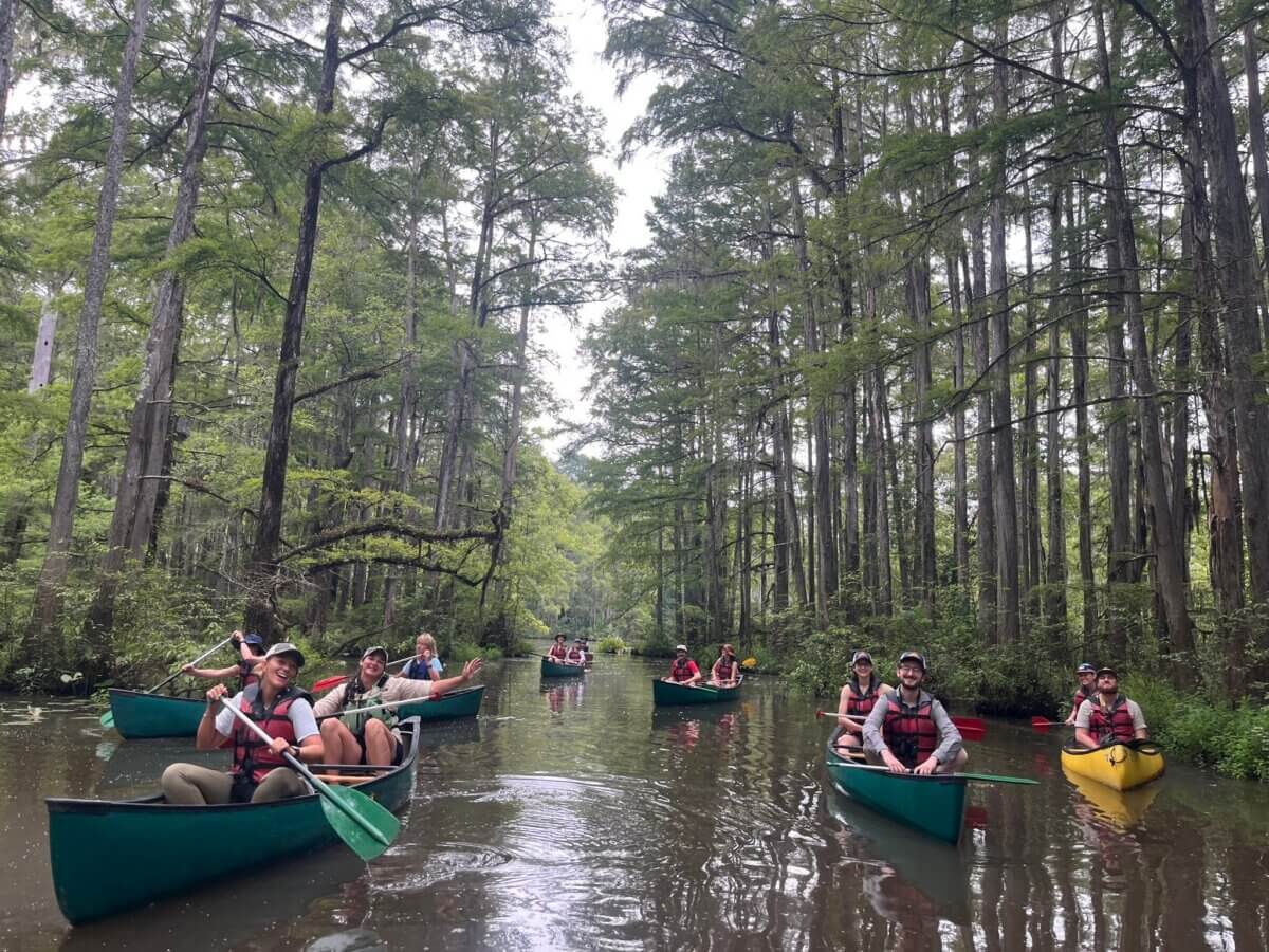 8 Ways to Get on the Water around BHAM, All Within an Hour’s Drive - 2 A group of people wearing life jackets paddle canoes on a calm, tree-lined river under an overcast sky along the Cahaba Blueway.