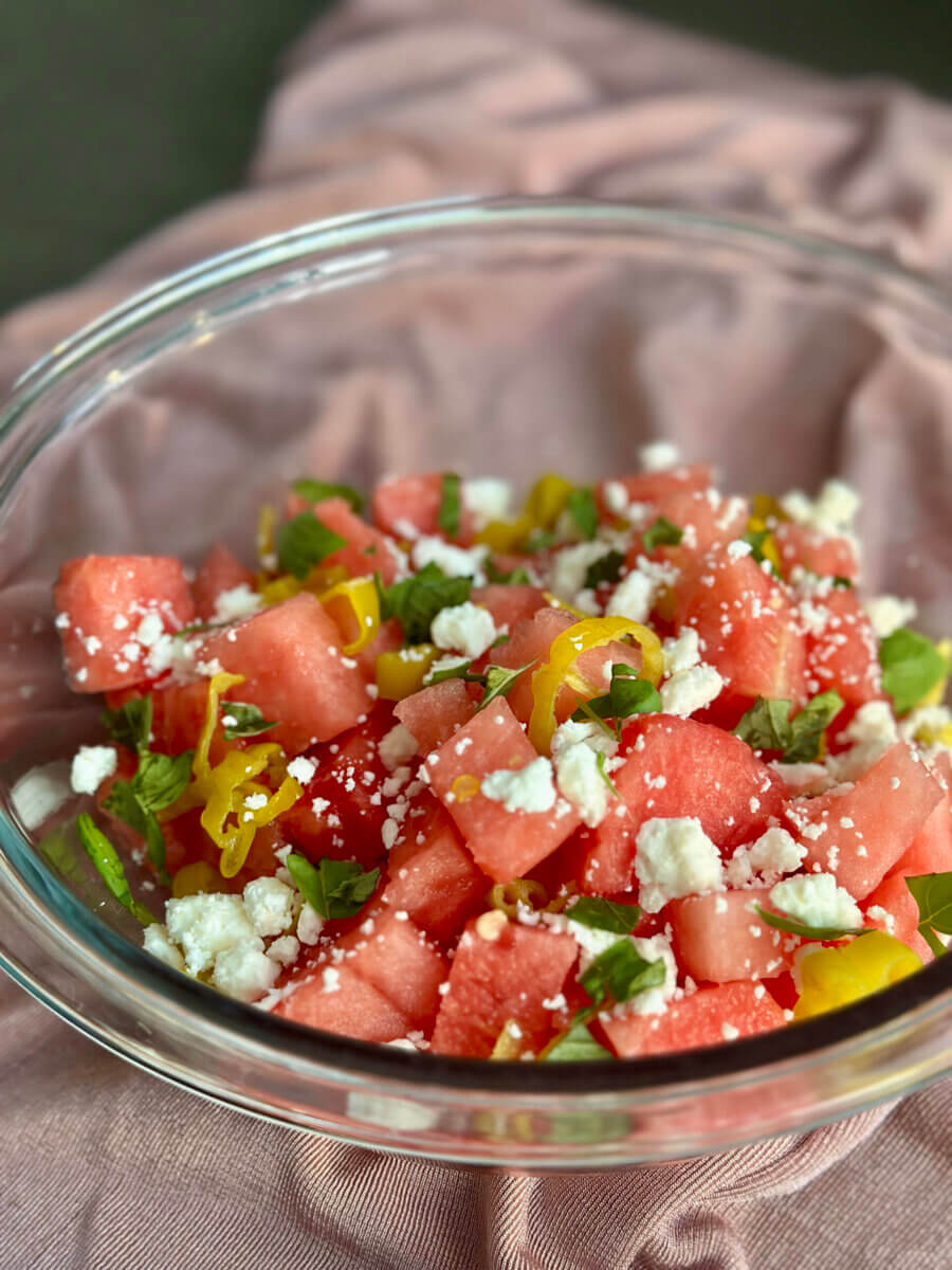 RECIPE: Watermelon Pepperoncini Salad - 2 A glass bowl filled with a watermelon salad topped with feta cheese, chopped herbs, and sliced yellow peppers, placed on a pink clothβperfect for summer recipes and refreshing summer weekends.