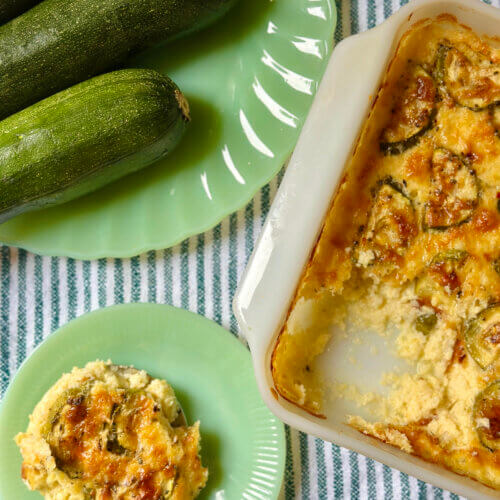 RECIPE: Zucchini Cheddar Grits Casserole - 3 A baked grits casserole with a creamy, golden top sits next to fresh zucchinis on a green plate and a serving on a separate plate, all on a striped tablecloth.