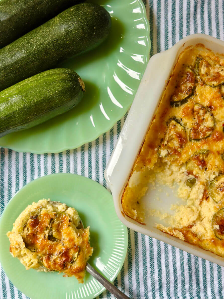 A baked grits casserole with a creamy, golden top sits next to fresh zucchinis on a green plate and a serving on a separate plate, all on a striped tablecloth.