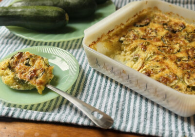 A baked zucchini casserole in a glass dish sits on a striped cloth, with a portion served on a green plate next to whole zucchinis.