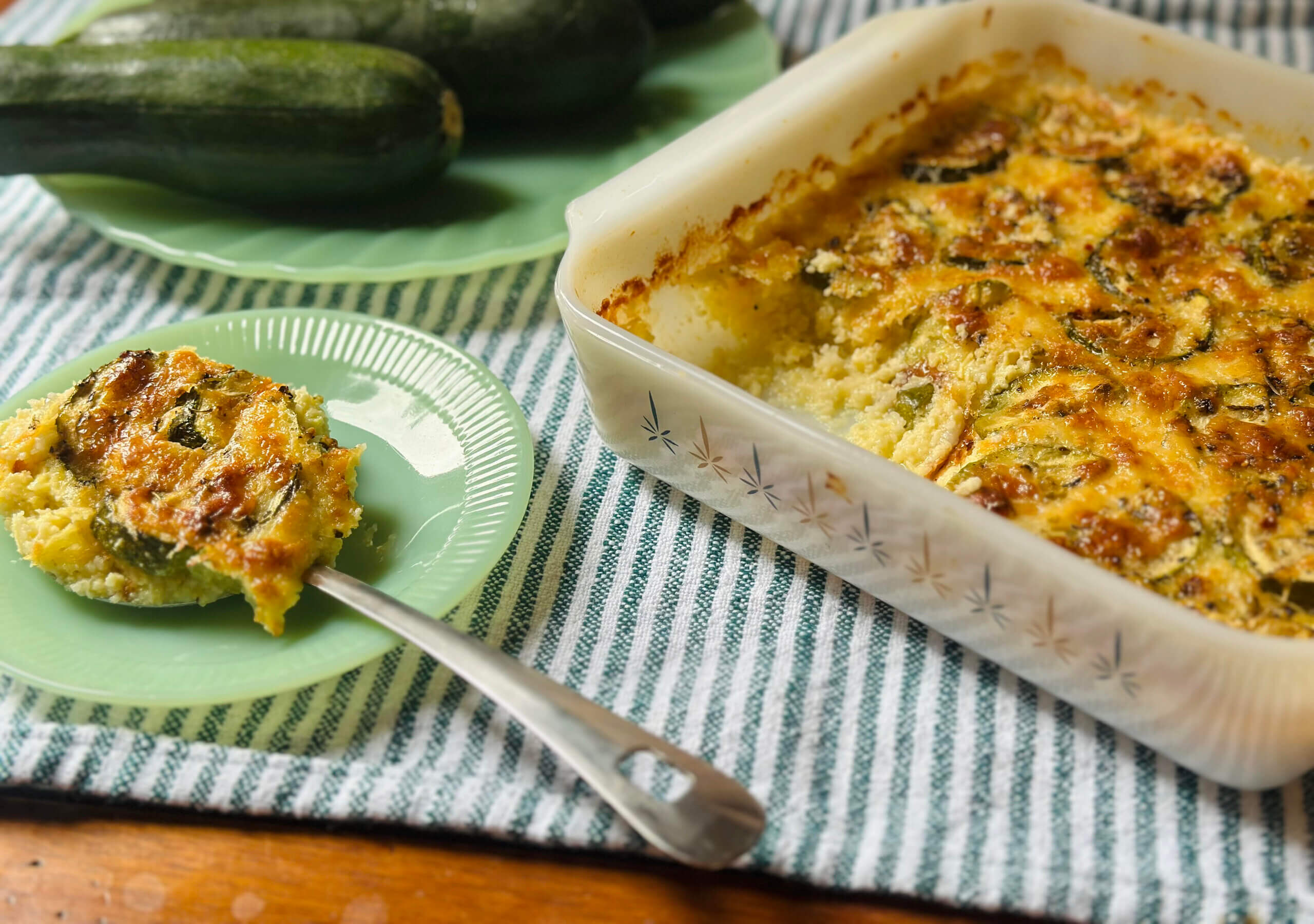 A baked zucchini casserole in a glass dish sits on a striped cloth, with a portion served on a green plate next to whole zucchinis.