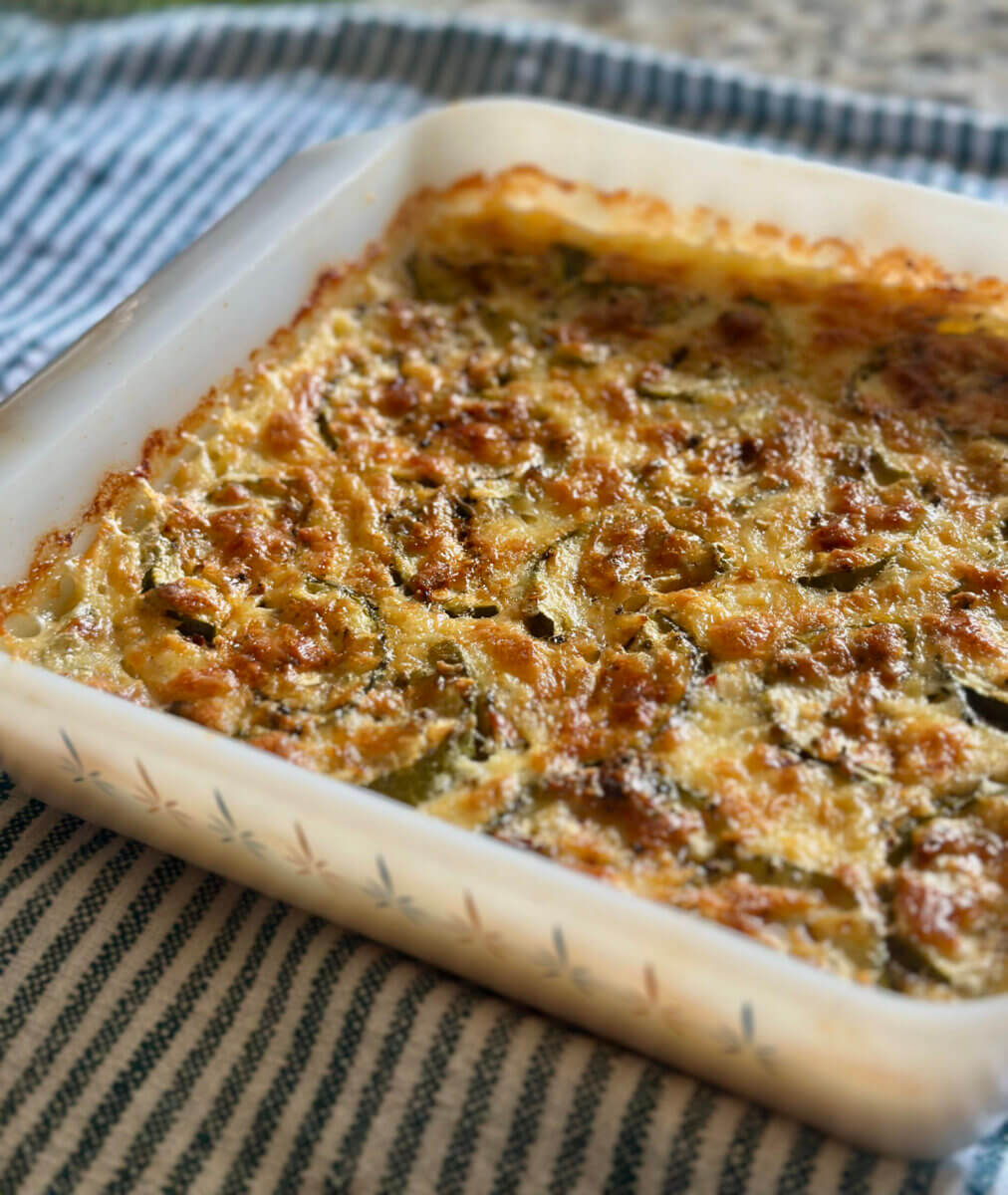 A baked casserole with a golden-brown, crispy top in a square white dish, resting on a striped cloth—perfect for summer weekend recipes.