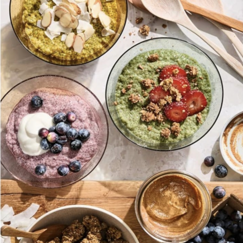 RECIPE: Chia Pudding 3 Ways - 3 Four bowls of colorful smoothie or porridge topped with fruit, coconut, and granola are arranged on a table with wooden spoons, a jar of nut butter, and loose blueberries.