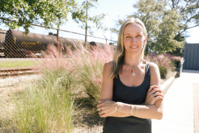 Jane Pope stands outdoors with arms crossed, wearing a black tank top and Jane Pope Jewelry, in front of pink grasses and a chain-link fence near a train track. Image by Lindsey Belle.