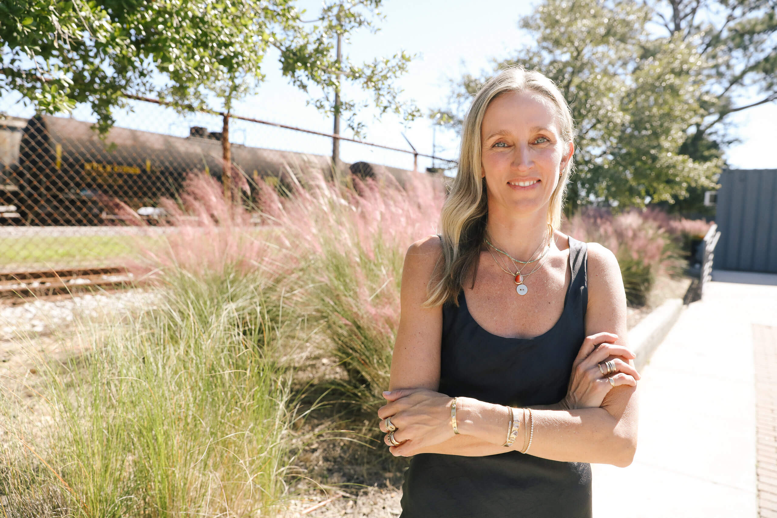 Jane Pope stands outdoors with arms crossed, wearing a black tank top and Jane Pope Jewelry, in front of pink grasses and a chain-link fence near a train track. Image by Lindsey Belle.
