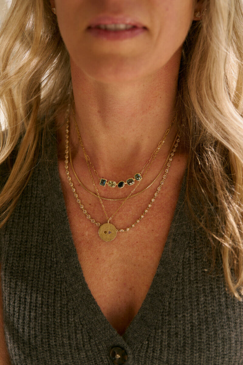 A person wearing a grey knit top and three layered gold necklaces from Jane Pope Jewelry, one featuring a round pendant and another adorned with green gemstones. Brand Spotlight on elevated everyday style.