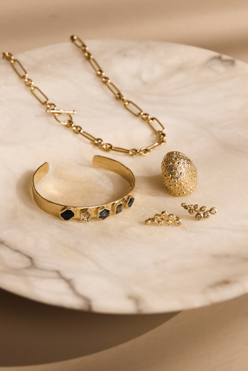 Brand Spotlight: Jane Pope Jewelry presents a gold chain necklace, a textured gold ring, a gold cuff bracelet with black stones, and two pairs of gold earrings displayed on a round marble plate.