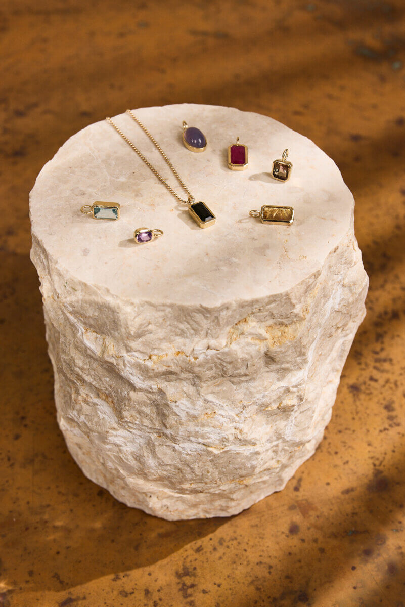 A collection of gold and gemstone pendants and rings from Jane Pope Jewelry is displayed on a rough, cylindrical stone pedestal against a brown background in this brand spotlight.