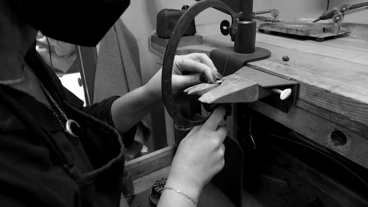 A person wearing a mask uses a saw to cut or shape a small object at a workbench in a workshop, showcasing the craftsmanship behind Jane Pope Jewelry in this Brand Spotlight.