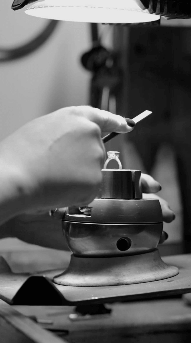 A person uses a tool to work on a ring held in a metal clamp under a desk lamp in the Jane Pope Jewelry workshop.