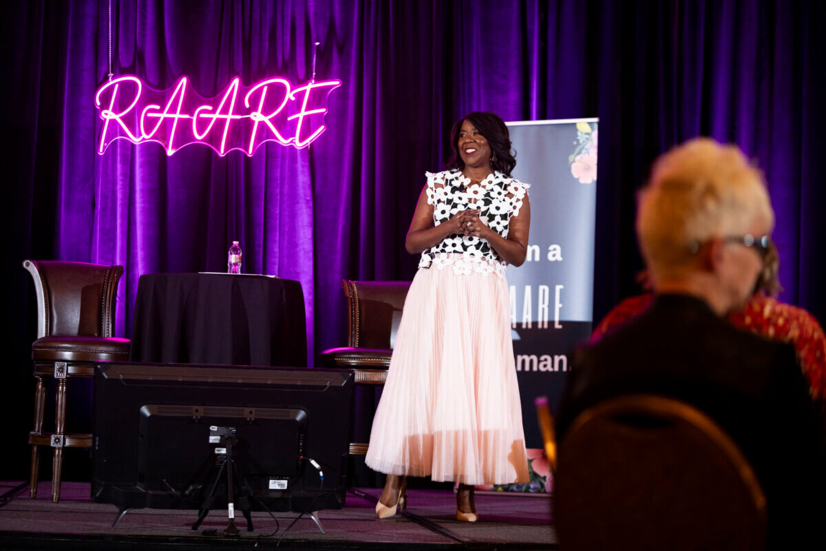 Meet a Woman Who's Reimagining the Modern Workplace - 4 A woman stands on stage smiling, wearing a white floral top and pleated skirt, with a neon "RAARE" sign and purple curtains in the background, as audience members watch from their seats.