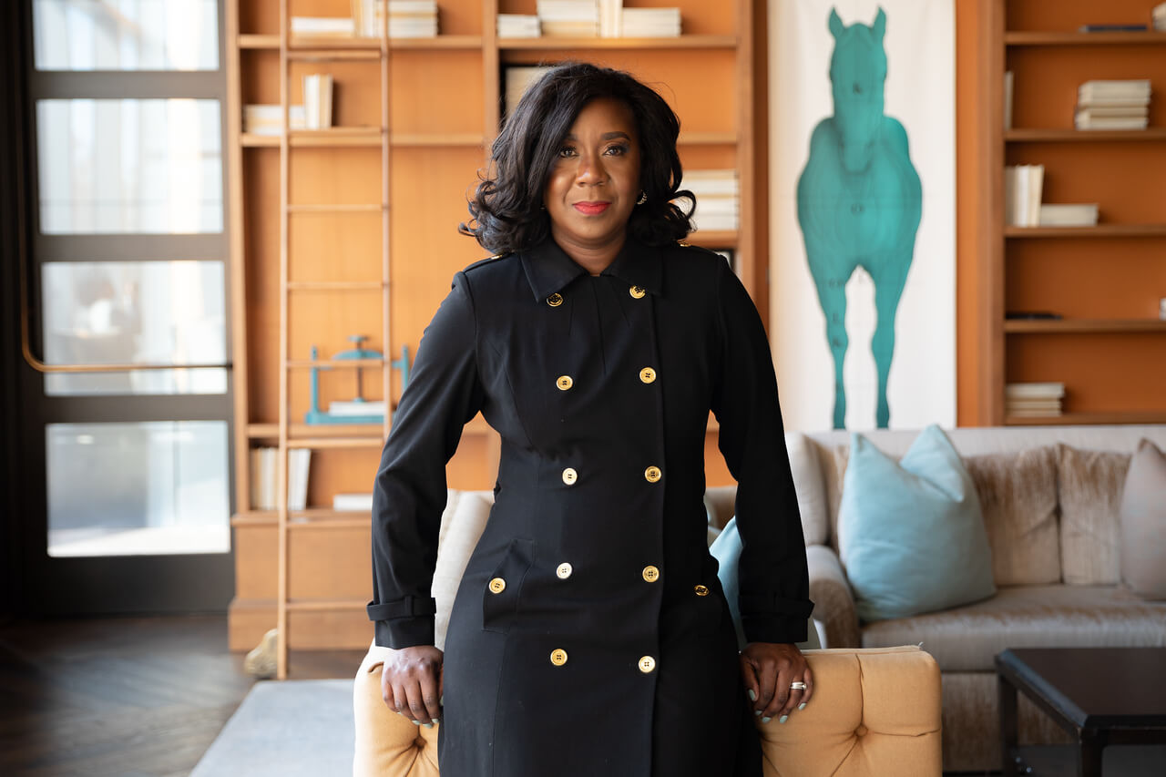 A woman in a black double-breasted coat stands before a beige sofa, building bridges to an inclusive future, with shelves and a large painting of a turquoise horse in the background.