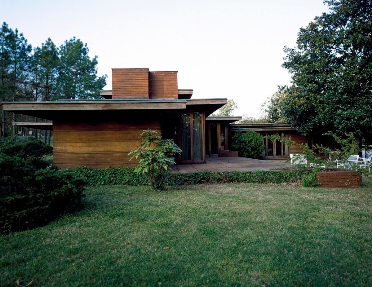 A modern, single-story house with flat roofs and a wooden exterior, by Frank Lloyd Wright, features large windows and surrounding greenery on a well-kept lawn—perfectly blending with Southern sites.
