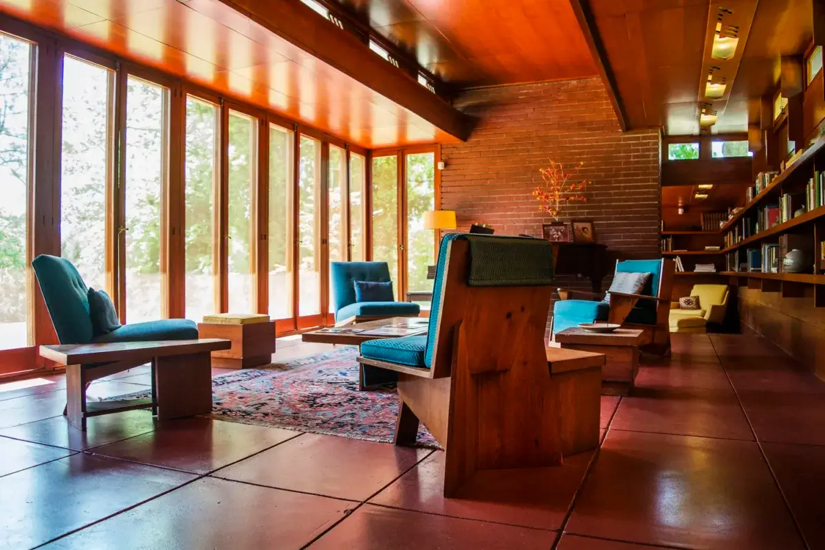 A modern living room with wooden furniture, blue upholstered chairs, large windows facing south, built-in bookshelves, and a patterned rug on a tiled floor.