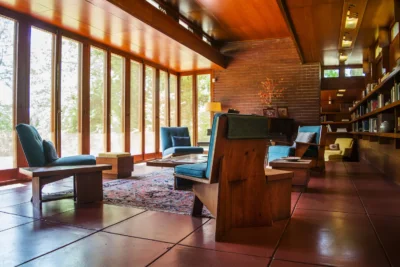 A modern living room with wooden furniture, blue upholstered chairs, large windows facing south, built-in bookshelves, and a patterned rug on a tiled floor.