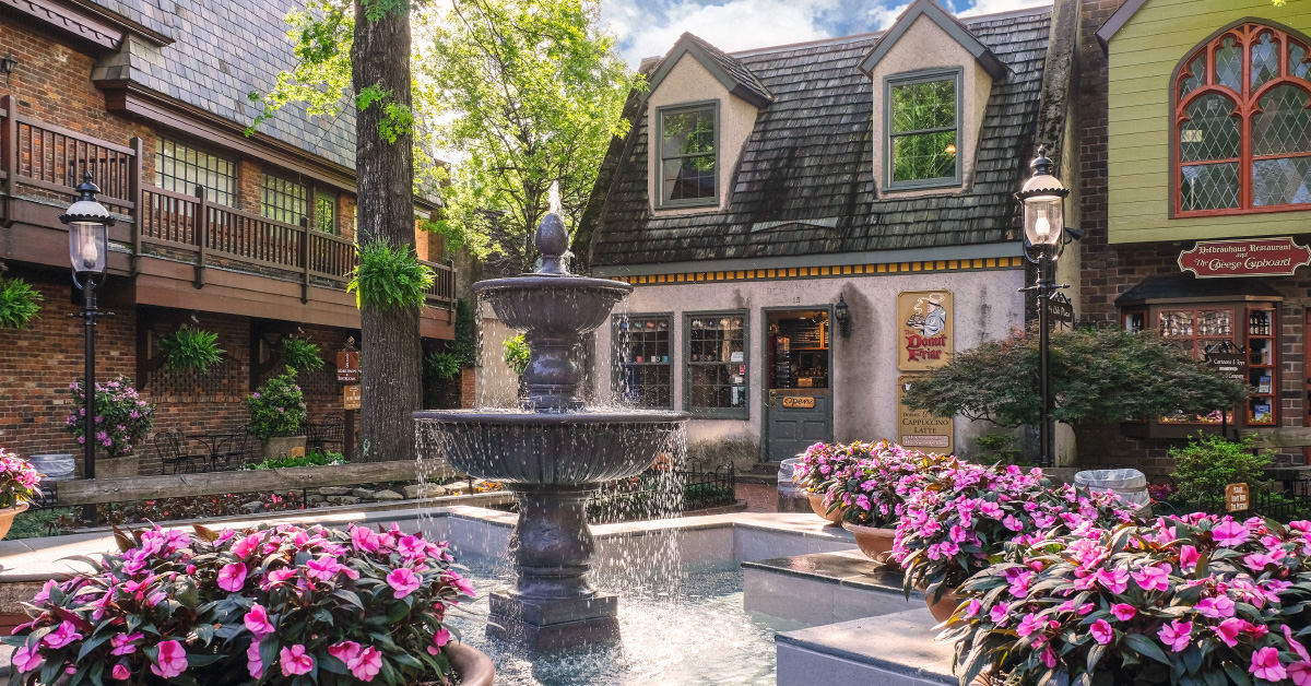 Where to Eat & Sip in Gatlinburg: From Local Favorites to Hidden Gems - 2 A stone fountain surrounded by pink flowers sits in a quaint courtyard with charming shopfronts, lush greenery, and cozy Gatlinburg restaurants under a blue sky.