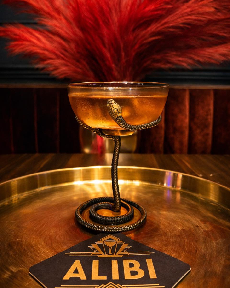 Where to Eat & Sip in Gatlinburg: From Local Favorites to Hidden Gems - 7 A cocktail in a glass with a snake-shaped stem sits on a round gold tray beside an “ALIBI” card, with red feathers blurred in the background.