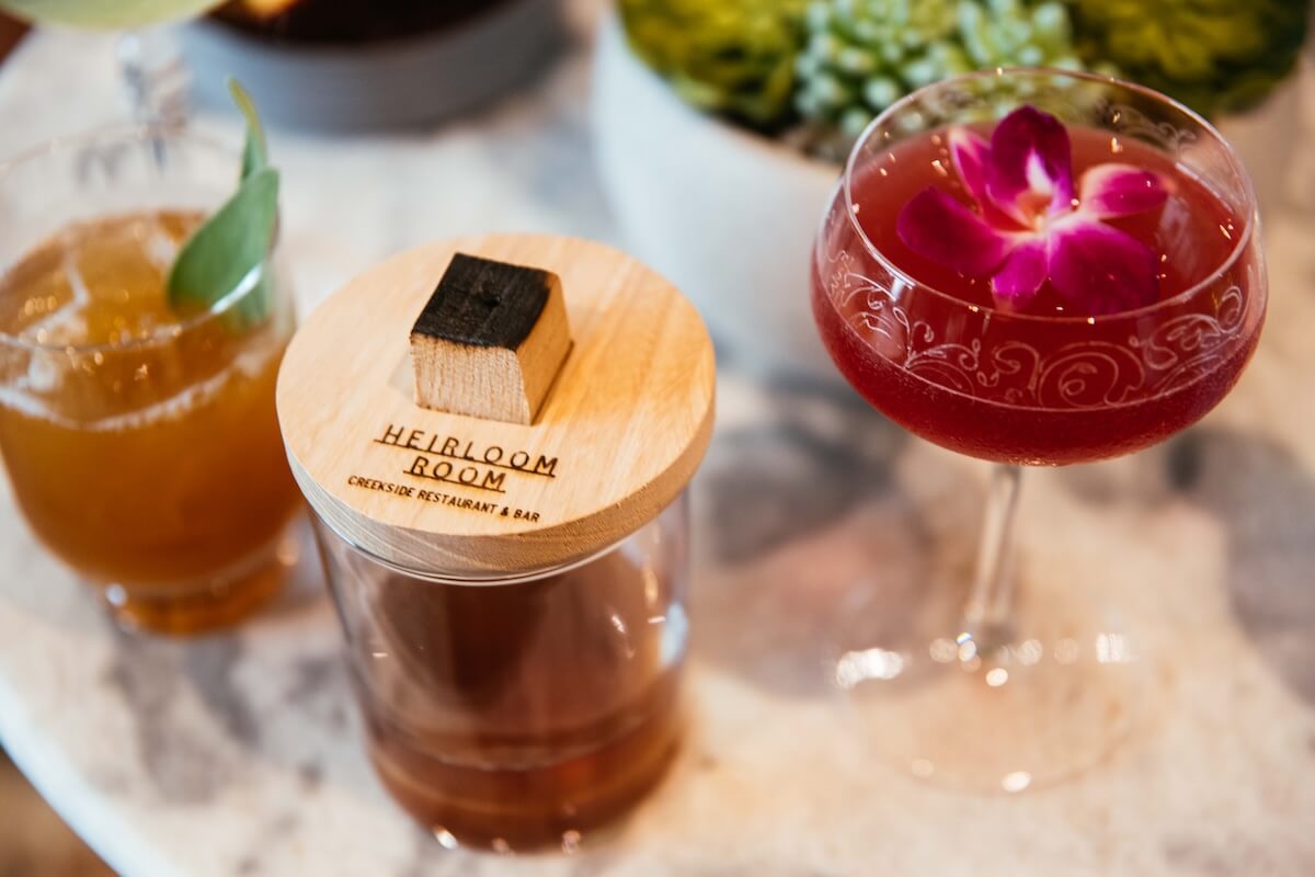 Where to Eat & Sip in Gatlinburg: From Local Favorites to Hidden Gems - 6 Three cocktails on a marble table: a local-inspired drink with an herbal garnish, one in a glass topped with a wooden lid labeled "Heirloom Room," and a pink cocktail garnished with a purple flower. Perfect for your Gatlinburg dining guide!.