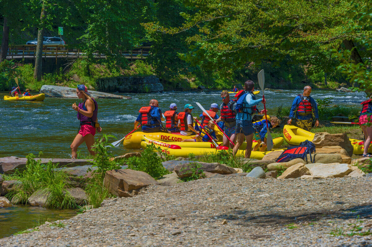 Great Southern Outdoors: Destinations for Active Travelers - 9 People wearing life vests prepare yellow rafts by a riverbank, while others stand nearby on rocks and gravel under trees on a sunny dayβan ideal scene for outdoor adventure vacations.