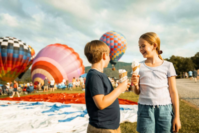Two children holding ice cream cones smile at each other in front of colorful hot air balloons and a crowd outdoors, capturing the joyful spirit of Southern Happenings during August 2026.