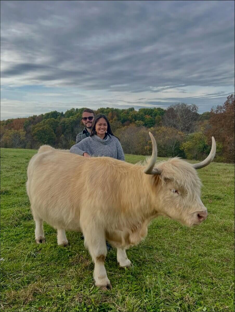 Two people stand smiling behind a fluffy cow in a grassy field with trees and a cloudy sky, capturing the charm of the Highland Trail.