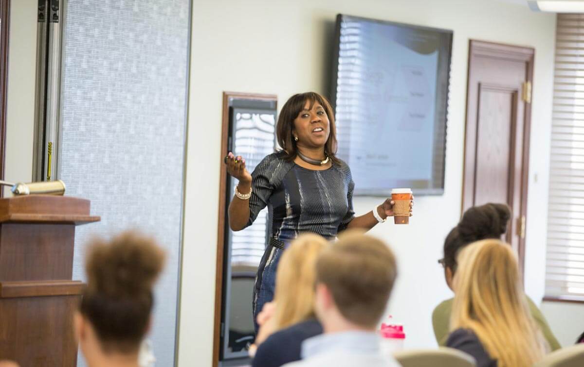 Meet a Woman Who's Reimagining the Modern Workplace - 3 A woman stands and speaks to a seated audience in a conference room, holding a cup.