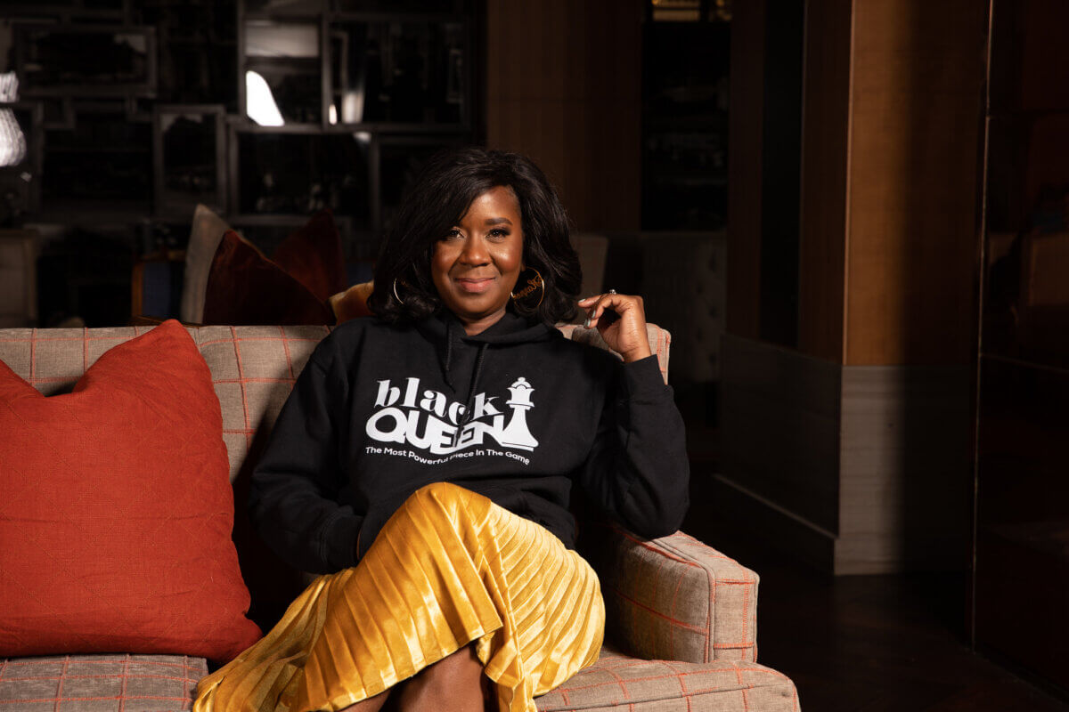 Meet a Woman Who's Reimagining the Modern Workplace - 6 A woman sits on a plaid sofa, wearing a black "Black Queen" hoodie and yellow pleated skirt, embodying strength and building bridges toward an inclusive future, with her legs crossed and hand resting near her face.