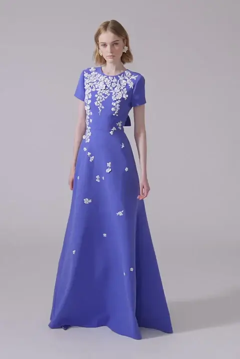 16 Luxe Louisville FINDS for July: Fine Art, Rare Bourbon & Resort-Ready Style - 8 A woman wearing a floor-length, short-sleeved, periwinkle blue gown with white floral appliqués on the bodice and upper skirt stands against a plain, light background—a Luxe Louisville look fit for July celebrations.