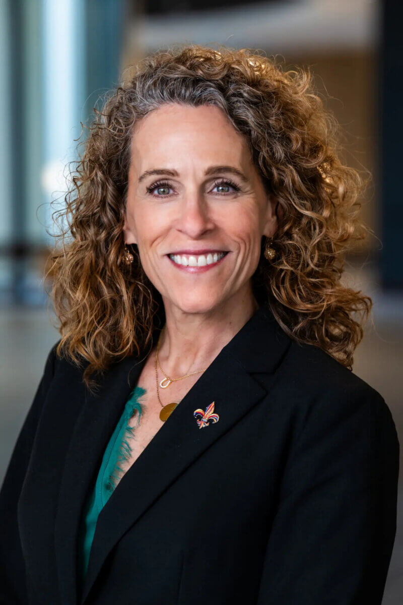 A woman with curly brown hair, known as Rebecca Fleischaker, wearing a black blazer, green top, and fleur-de-lis pin, smiles.