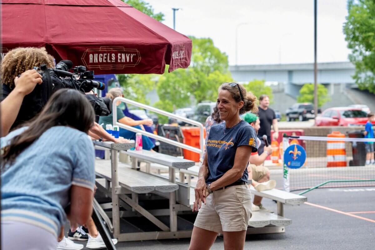 A woman stands outside smiling as she is interviewed by news crew under a large red umbrella, with people and traffic cones in the background.