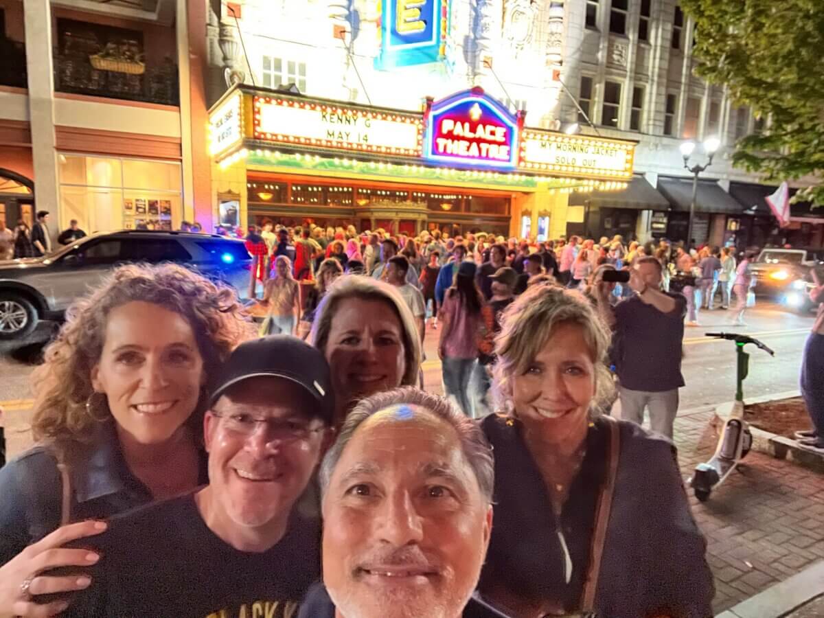 Five adults smile for a selfie at night outside the brightly lit Palace Theatre, with a large crowd gathered in front of the marquee.