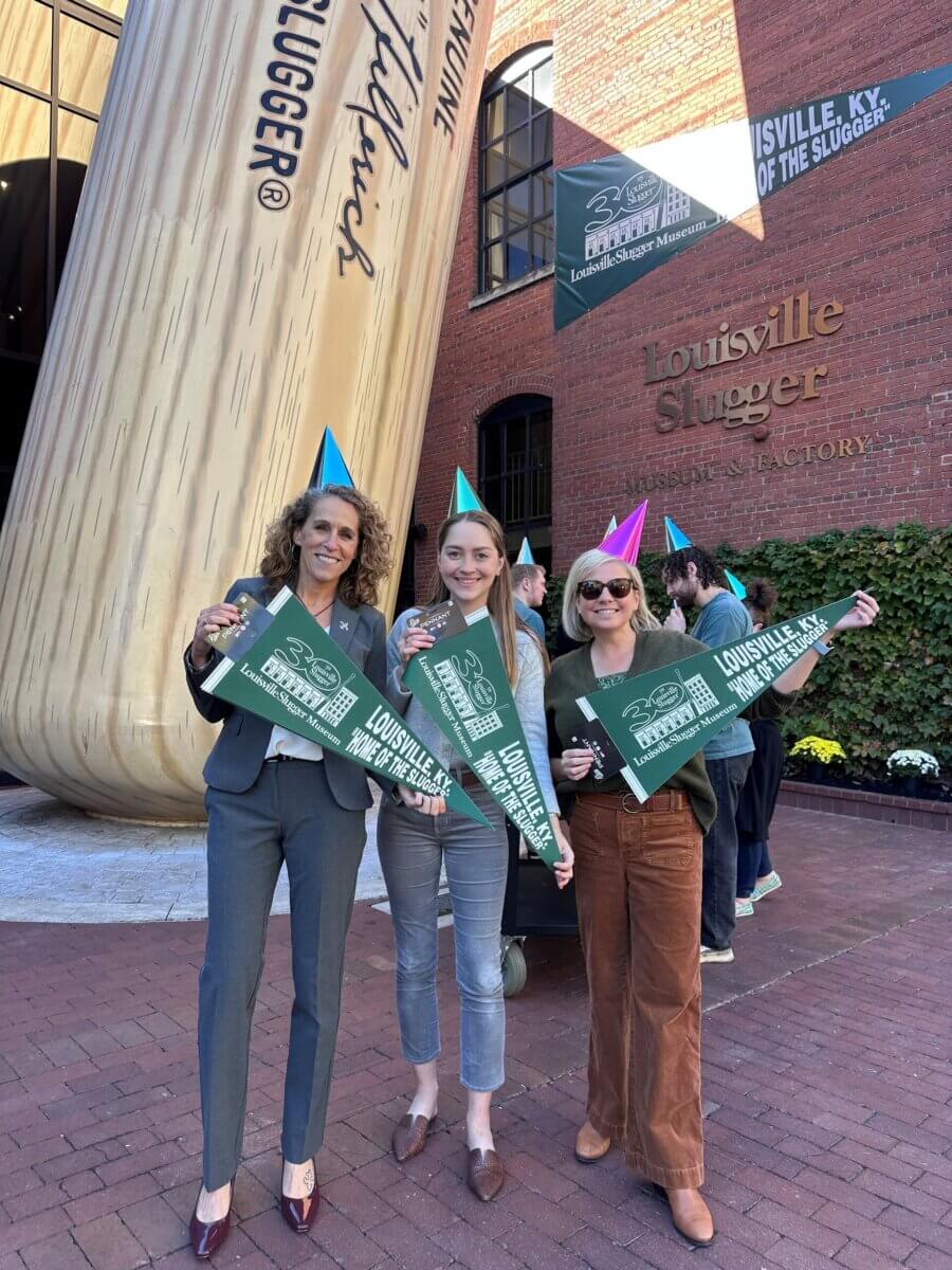 Three women wearing party hats hold Louisville Slugger signs and pennants, standing in front of the Louisville Slugger Museum & Factory and a giant baseball bat.
